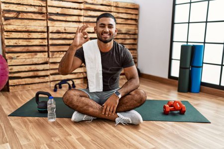 Photo pour Jeune homme indien assis sur tapis d'entraînement à la salle de gym souriant positif faire ok signe avec la main et les doigts. expression réussie. - image libre de droit