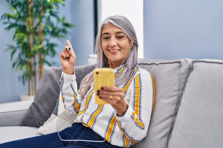 Foto de Mujer de mediana edad con el pelo gris usando el teléfono inteligente sentado en el sofá sonriendo feliz señalando con la mano y el dedo a un lado - Imagen libre de derechos