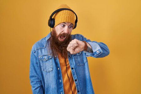 Téléchargez les photos : Homme caucasien à longue barbe écoutant de la musique à l'aide d'écouteurs regardant le temps de la montre inquiet, peur de se retrouver en retard - en image libre de droit