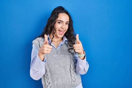 Photo for Young brunette woman standing over blue background pointing fingers to camera with happy and funny face. good energy and vibes. - Royalty Free Image