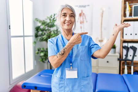 Photo for Middle age grey-haired woman wearing physiotherapist uniform at medical clinic amazed and smiling to the camera while presenting with hand and pointing with finger. - Royalty Free Image
