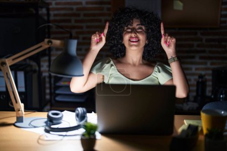 Photo for Young brunette woman with curly hair working at the office at night smiling amazed and surprised and pointing up with fingers and raised arms. - Royalty Free Image