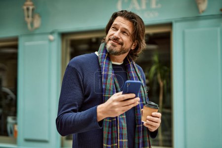 Téléchargez les photos : Moyen âge bel homme en utilisant smartphone boire un café à la ville - en image libre de droit