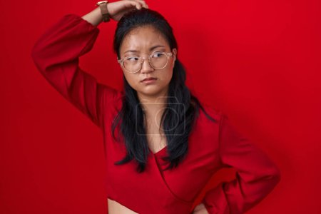 Téléchargez les photos : Asian young woman standing over red background confuse and wondering about question. uncertain with doubt, thinking with hand on head. pensive concept. - en image libre de droit