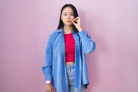 Téléchargez les photos : Jeune femme asiatique debout sur fond rose bouche et lèvres fermées comme zip avec les doigts. secret et silencieux, tabou parlant - en image libre de droit