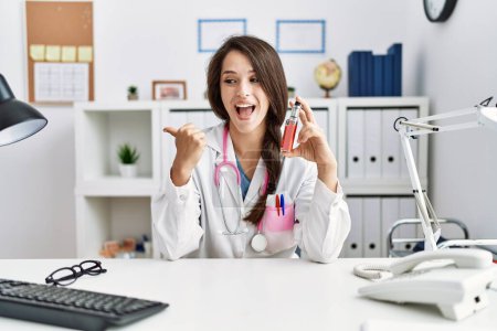 Photo for Young doctor woman holding electronic cigarette at medical clinic pointing thumb up to the side smiling happy with open mouth - Royalty Free Image