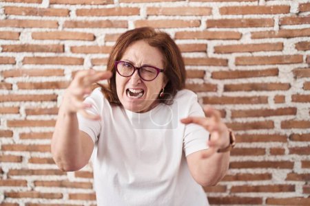 Foto de Senior woman with glasses standing over bricks wall shouting frustrated with rage, hands trying to strangle, yelling mad - Imagen libre de derechos