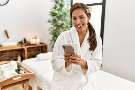 Photo for Young latin woman wearing bathrobe using smartphone - Royalty Free Image