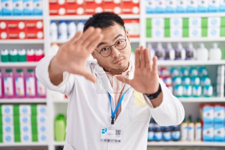 Photo for Chinese young man working at pharmacy drugstore doing frame using hands palms and fingers, camera perspective - Royalty Free Image