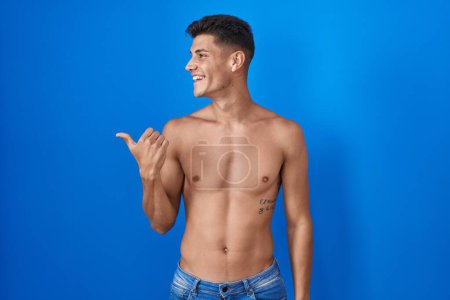 Foto de Joven hombre hispano de pie sin camisa sobre fondo azul sonriendo con la cara feliz mirando y señalando a un lado con el pulgar hacia arriba. - Imagen libre de derechos