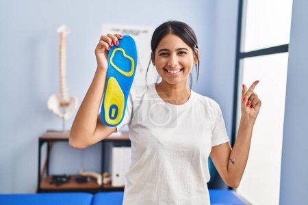 Téléchargez les photos : Jeune femme hispanique tenant semelle intérieure de chaussure à la clinique de physiothérapie souriant heureux pointant avec la main et le doigt vers le côté - en image libre de droit