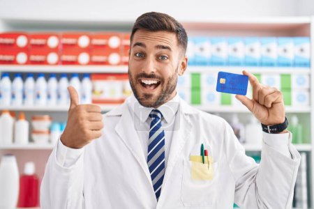Téléchargez les photos : Bel homme hispanique travaillant à la pharmacie pharmacie tenant carte de crédit souriant heureux et positif, pouce levé faire excellent et signe d'approbation - en image libre de droit