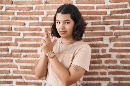 Foto de Young hispanic woman standing over bricks wall holding symbolic gun with hand gesture, playing killing shooting weapons, angry face - Imagen libre de derechos