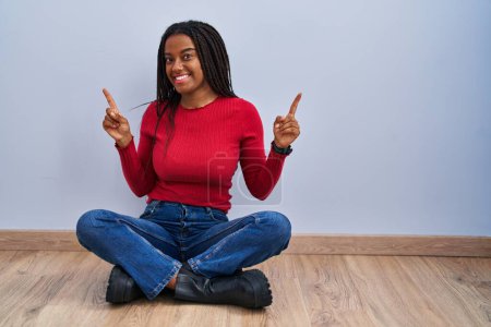 Photo for Young african american with braids sitting on the floor at home smiling confident pointing with fingers to different directions. copy space for advertisement - Royalty Free Image