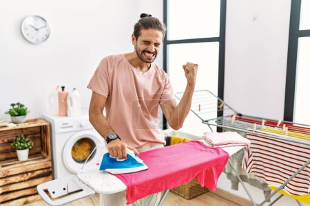Foto de Joven hombre hispano planchando ropa en casa muy feliz y emocionado haciendo gesto ganador con los brazos levantados, sonriendo y gritando por el éxito. concepto de celebración. - Imagen libre de derechos