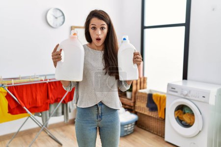 Photo for Young brunette woman holding detergent bottle at laundry room in shock face, looking skeptical and sarcastic, surprised with open mouth - Royalty Free Image