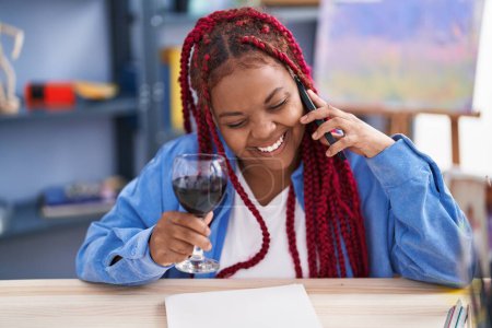 Téléchargez les photos : African american woman artist talking on smartphone drinking wine at art studio - en image libre de droit