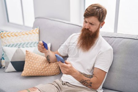 Téléchargez les photos : Young redhead man using smartphone and credit card sitting on sofa at home - en image libre de droit