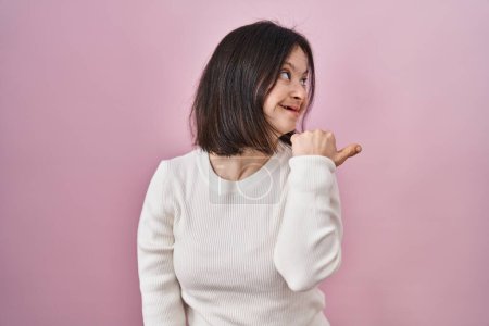 Photo for Woman with down syndrome standing over pink background smiling with happy face looking and pointing to the side with thumb up. - Royalty Free Image