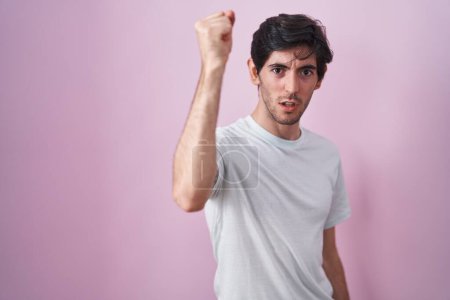Foto de Young hispanic man standing over pink background angry and mad raising fist frustrated and furious while shouting with anger. rage and aggressive concept. - Imagen libre de derechos