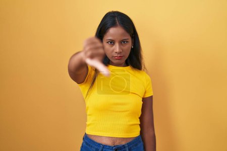 Téléchargez les photos : Jeune femme indienne debout sur fond jaune regardant malheureux et en colère montrant rejet et négatif avec les pouces vers le bas geste. mauvaise expression. - en image libre de droit