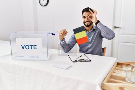 Foto de Joven hombre guapo con barba en las elecciones de campaña política sosteniendo la bandera de Alemania sonriendo feliz haciendo señal bien con la mano en el ojo mirando a través de los dedos - Imagen libre de derechos