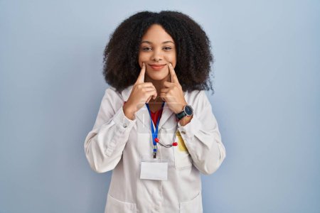 Téléchargez les photos : Jeune femme afro-américaine portant l'uniforme de médecin et stéthoscope souriant avec la bouche ouverte, les doigts pointant et forçant sourire joyeux - en image libre de droit