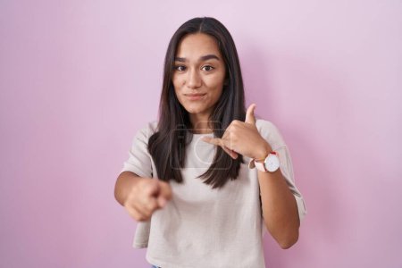 Photo for Young hispanic woman standing over pink background smiling doing talking on the telephone gesture and pointing to you. call me. - Royalty Free Image