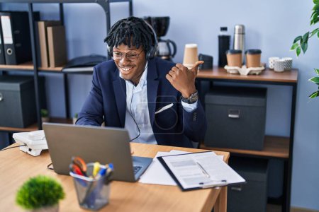 Téléchargez les photos : Jeune homme africain avec dreadlocks travaillant au bureau portant casque pointant pouce vers le côté souriant heureux avec la bouche ouverte - en image libre de droit