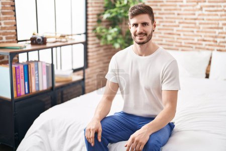 Téléchargez les photos : Jeune homme caucasien souriant confiant assis sur le lit dans la chambre - en image libre de droit