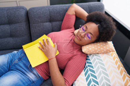 Foto de Mujer afroamericana sosteniendo libro acostada en sofá durmiendo en casa - Imagen libre de derechos