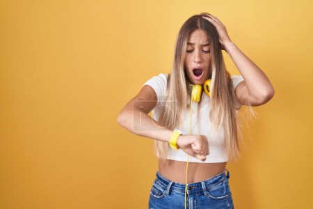 Téléchargez les photos : Young blonde woman standing over yellow background wearing headphones looking at the watch time worried, afraid of getting late - en image libre de droit