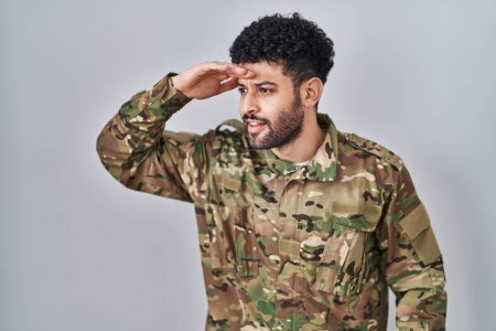 Photo for Arab man wearing camouflage army uniform very happy and smiling looking far away with hand over head. searching concept. - Royalty Free Image