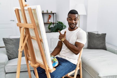 Téléchargez les photos : Jeune homme africain peignant sur toile à la maison gai avec un sourire sur le visage pointant avec la main et le doigt sur le côté avec une expression heureuse et naturelle - en image libre de droit
