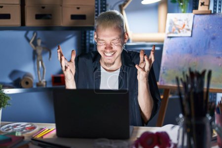 Téléchargez les photos : Jeune homme caucasien utilisant ordinateur portable la nuit au studio d'art célébrant fou et fou de succès avec les bras levés et les yeux fermés hurlant excité. concept gagnant - en image libre de droit