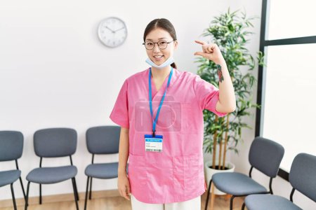 Photo for Young asian nurse woman at medical waiting room smiling and confident gesturing with hand doing small size sign with fingers looking and the camera. measure concept. - Royalty Free Image