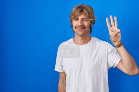 Téléchargez les photos : Homme d'âge moyen debout sur fond bleu montrant et pointant vers le haut avec les doigts numéro trois tout en souriant confiant et heureux. - en image libre de droit
