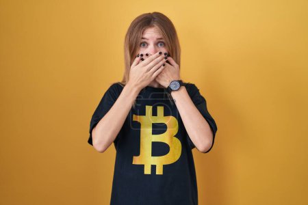 Foto de Mujer rubia caucásica con camiseta bitcoin impactada cubriendo la boca con las manos por error. concepto secreto. - Imagen libre de derechos