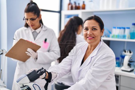 Photo for Three woman scientists using microscope write on checklist at laboratory - Royalty Free Image