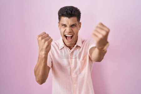 Téléchargez les photos : Jeune homme hispanique debout sur fond rose en colère et fou levant les poings frustrés et furieux tout en criant de colère. rage et concept agressif. - en image libre de droit