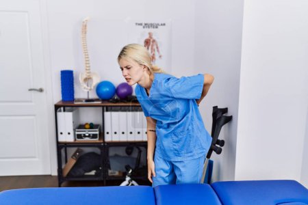 Photo for Young physiotherapist woman working at pain recovery clinic suffering of backache, touching back with hand, muscular pain - Royalty Free Image