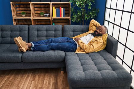 Téléchargez les photos : Jeune homme afro-américain souriant confiant détendu couché sur le canapé à la maison - en image libre de droit