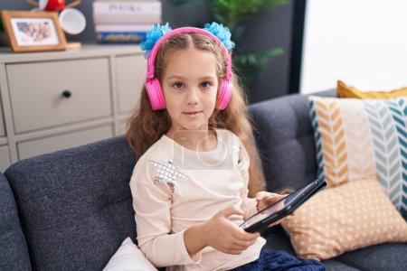 Téléchargez les photos : Adorable caucasian girl using touchpad and headphones sitting on sofa at home - en image libre de droit