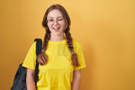Téléchargez les photos : Jeune femme caucasienne portant sac à dos étudiant sur fond jaune clin d'oeil en regardant la caméra avec une expression sexy, gai et heureux visage. - en image libre de droit