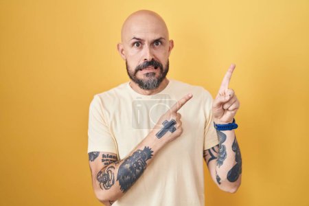 Foto de Hombre hispano con tatuajes de pie sobre fondo amarillo señalando a un lado preocupado y nervioso con ambas manos, preocupado y sorprendido expresión - Imagen libre de derechos