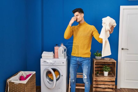 Téléchargez les photos : Jeune homme hispanique laver les vêtements sentant les vêtements sales à la blanchisserie - en image libre de droit