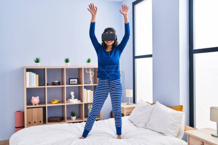 Photo for Young latin woman using virtual reality glasses standing on bed at bedroom - Royalty Free Image