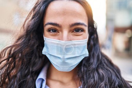 Téléchargez les photos : Jeune femme hispanique souriante confiante portant un masque médical dans la rue - en image libre de droit