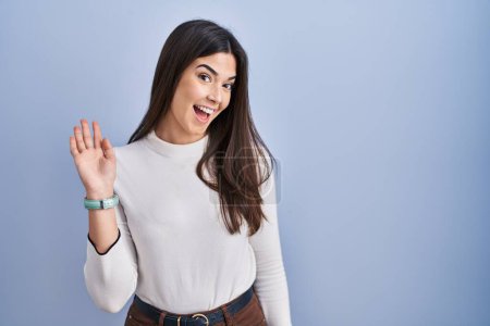 Téléchargez les photos : Jeune femme brune debout sur fond bleu renonçant à dire bonjour heureux et souriant, geste de bienvenue amical - en image libre de droit