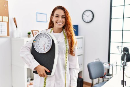 Téléchargez les photos : Jeune femme rousse nutritionniste médecin tenant la machine de pesage regardant positif et heureux debout et souriant avec un sourire confiant montrant les dents - en image libre de droit
